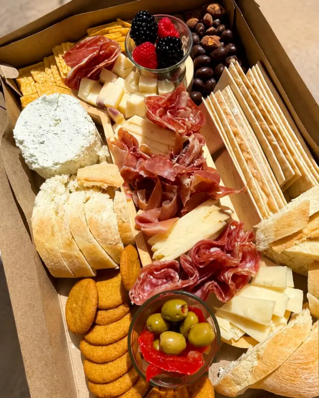 Close overhead of a boxed board: prosciutto, cubed cheese, sliced baguette, breadsticks, crackers, olives and berries