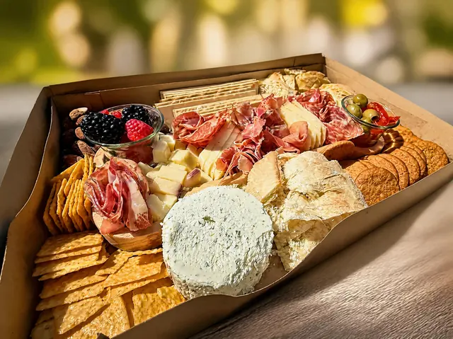 A boxed board packed edge to edge with cured meats, a whole cheese round, bread and crackers, on a table outdoors