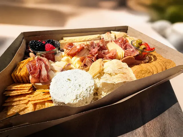 An open kraft box of cured meats, a whole cheese round, bread and crackers, on a dark wood table in the sun