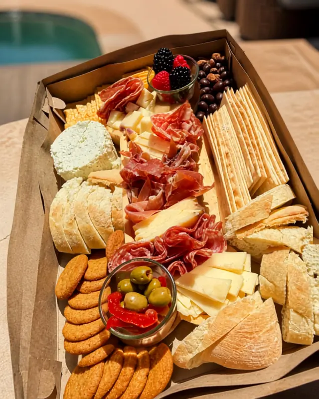 A boxed charcuterie board from above on a poolside table — prosciutto, cheeses, sliced bread, breadsticks, crackers and olives